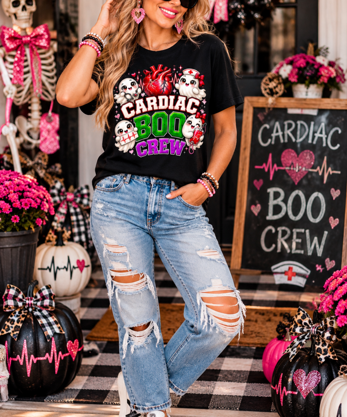 Woman standing in black Cardiac Boo Crew cardiology nurse Halloween T-shirt with ripped jeans, pink Halloween pumpkins and festive porch decorations.