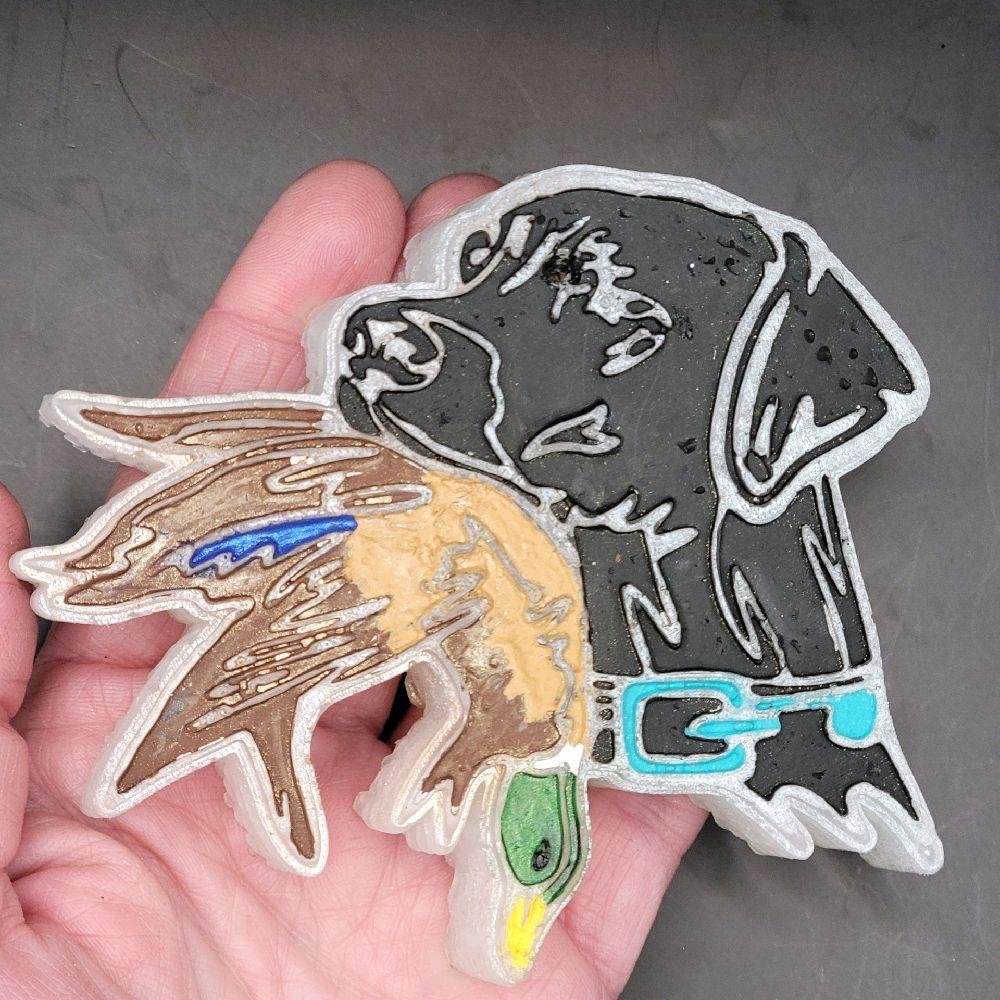 Black Lab holding a duck car freshie resting in a hand to show its relative size.